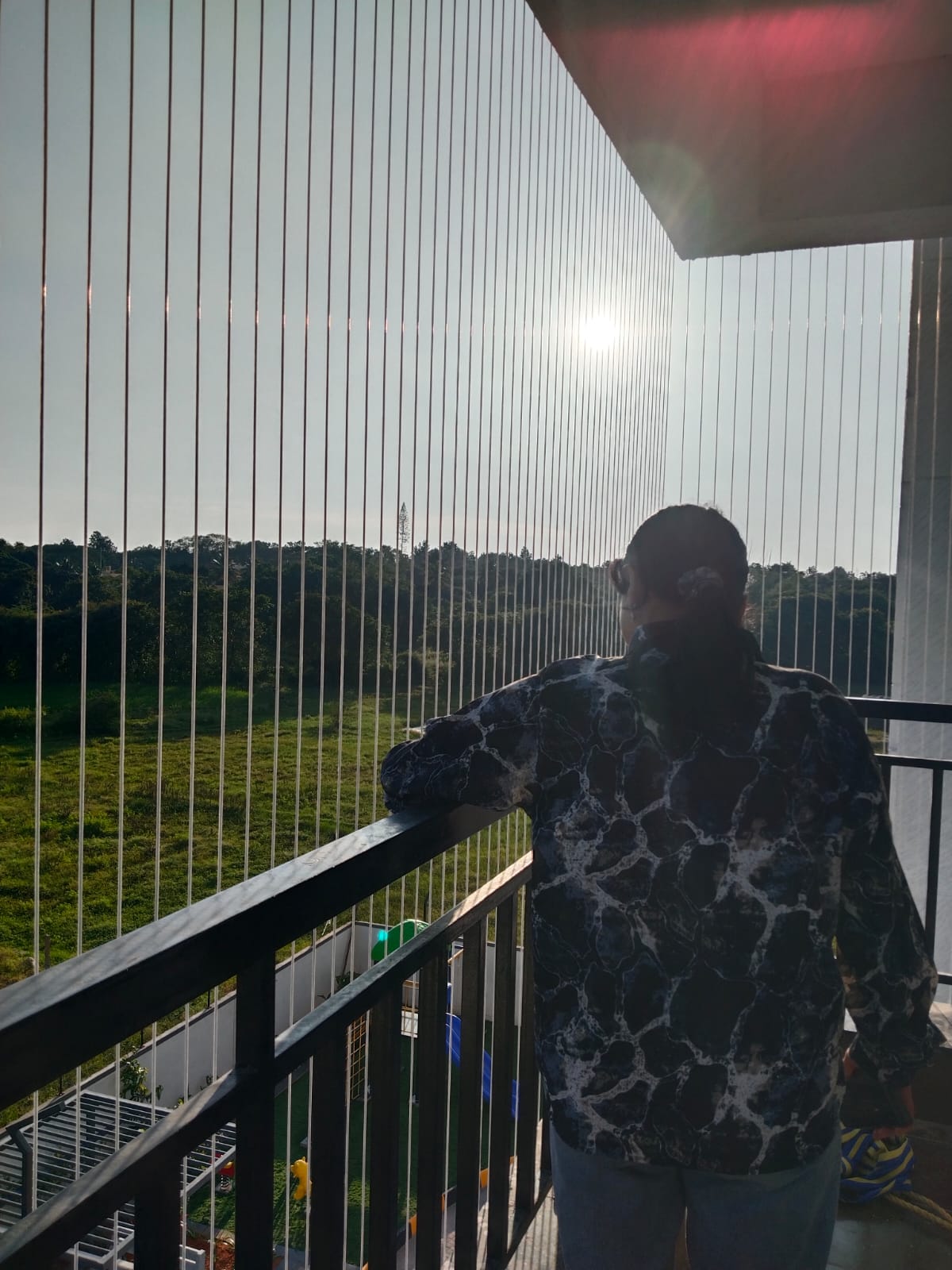 L-shape apartment balcony in Bengaluru fitted with PR invisible grill cables, with a resident leaning on the railing looking out at green fields