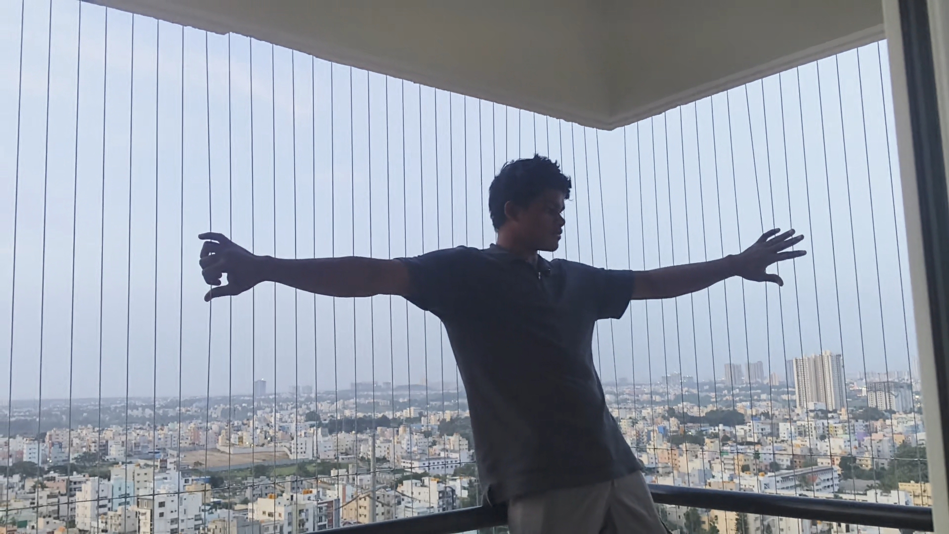 U-shape high-rise balcony in Bengaluru covered with PR invisible grill cables across three sides, with a man standing arms outstretched in front of the city skyline